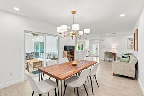 Un espace repas moderne avec une table en bois et des chaises blanches, un lustre, et une vue sur un salon comprenant un canapé et un téléviseur.