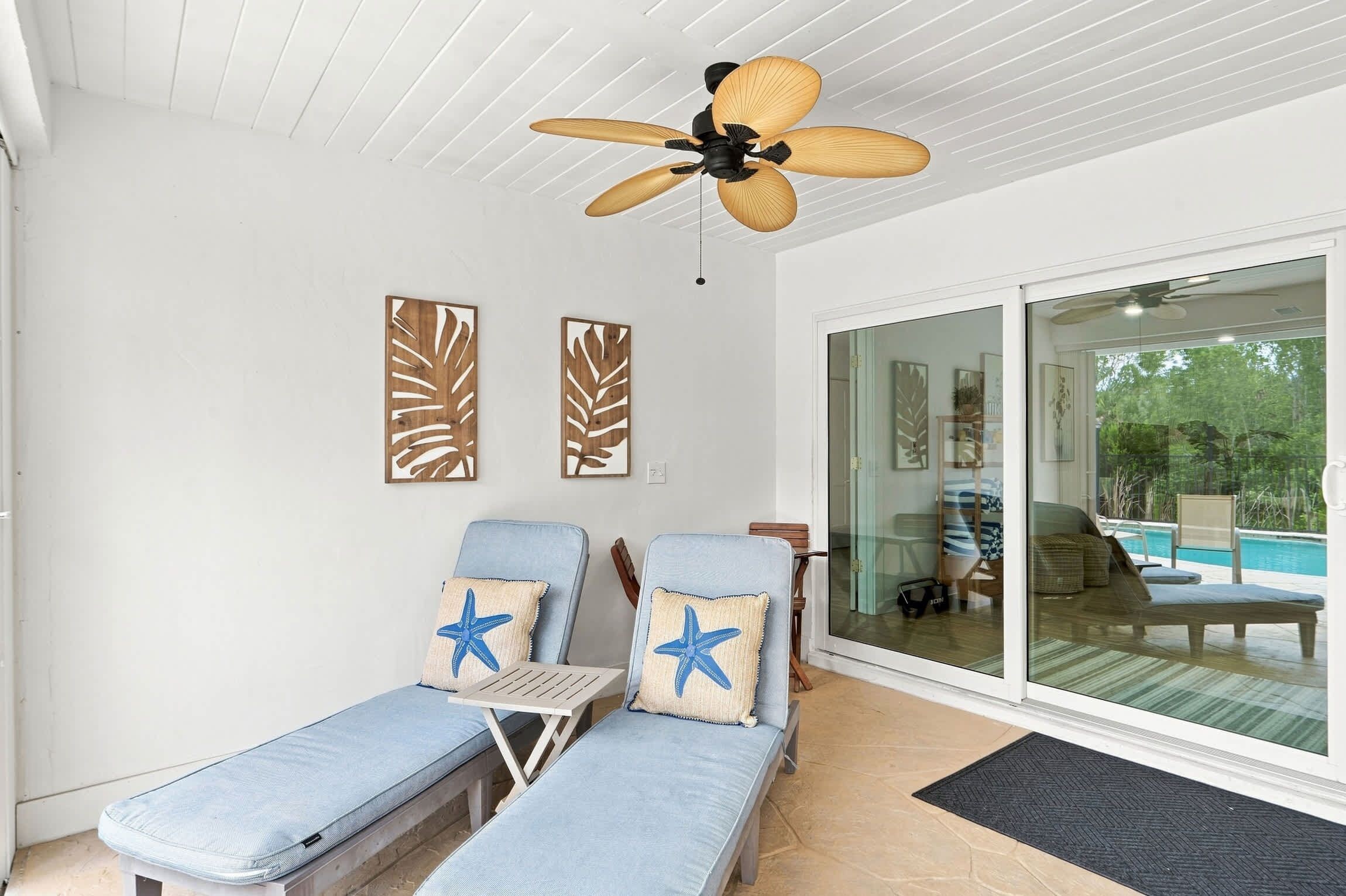 Une pièce baignée de lumière, dotée d’un ventilateur de plafond, de deux fauteuils confortables ornés de coussins en forme d’étoile de mer, et d’une porte vitrée donnant sur un espace piscine.