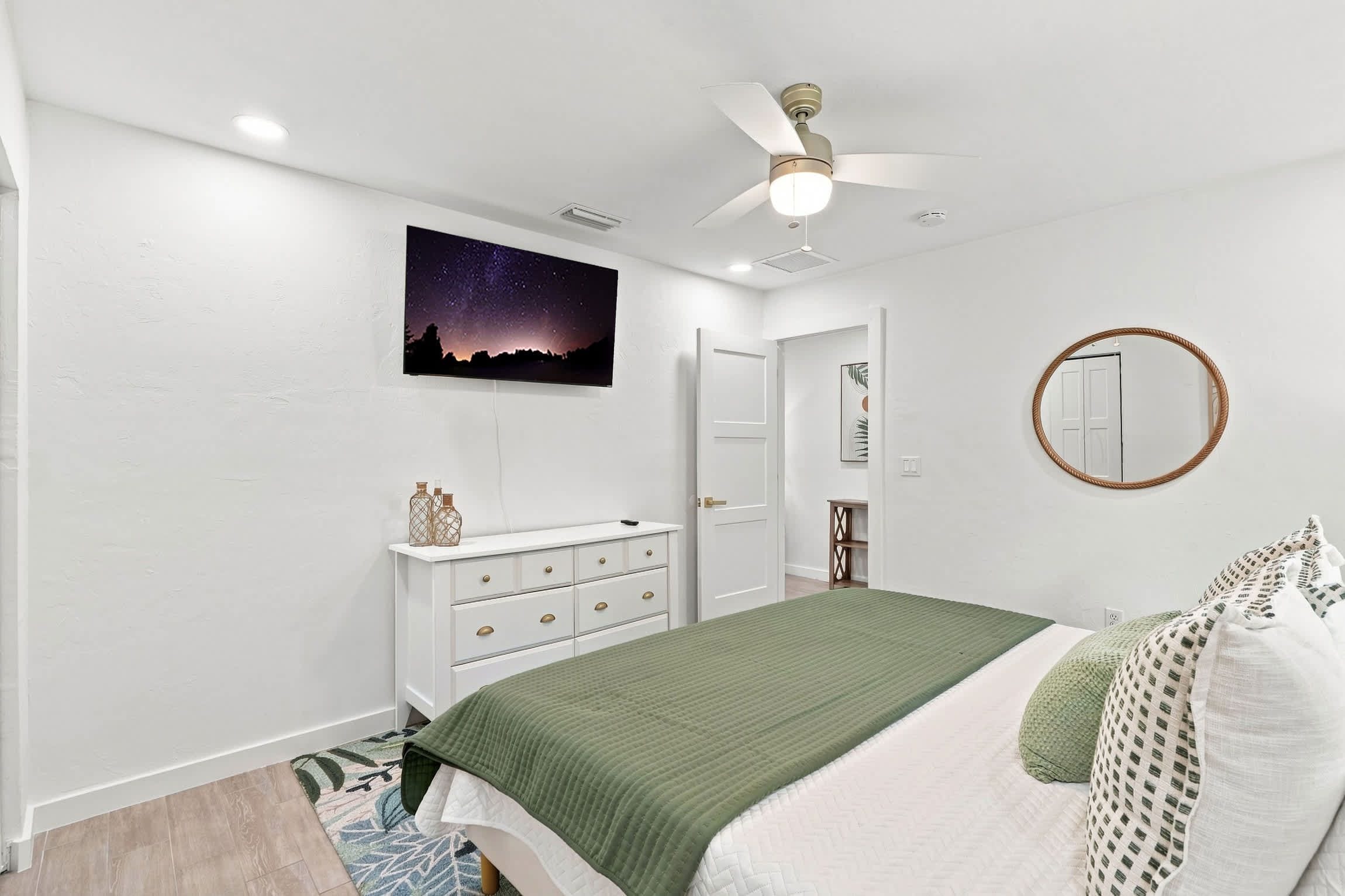 A bedroom with a bed, dresser, ceiling fan, night sky artwork, and a round mirror.