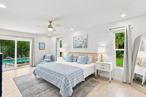 Une chambre à coucher avec un lit, des tables de chevet, un ventilateur de plafond, un tableau et une vue sur une piscine.