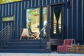 A modern building with a dark exterior, a set of stairs leading to a covered area with two wooden chairs and a red chair, and a glass door reflecting trees. - The Luna Cottage at Sandblox Resort (Benton Harbor)