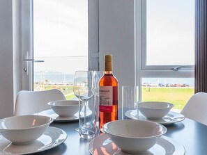 Ein Esstisch mit weißen Schüsseln, Weingläsern und einer Weinflasche, vor einem Fenster mit Blick auf eine Wiese und Gebäude.