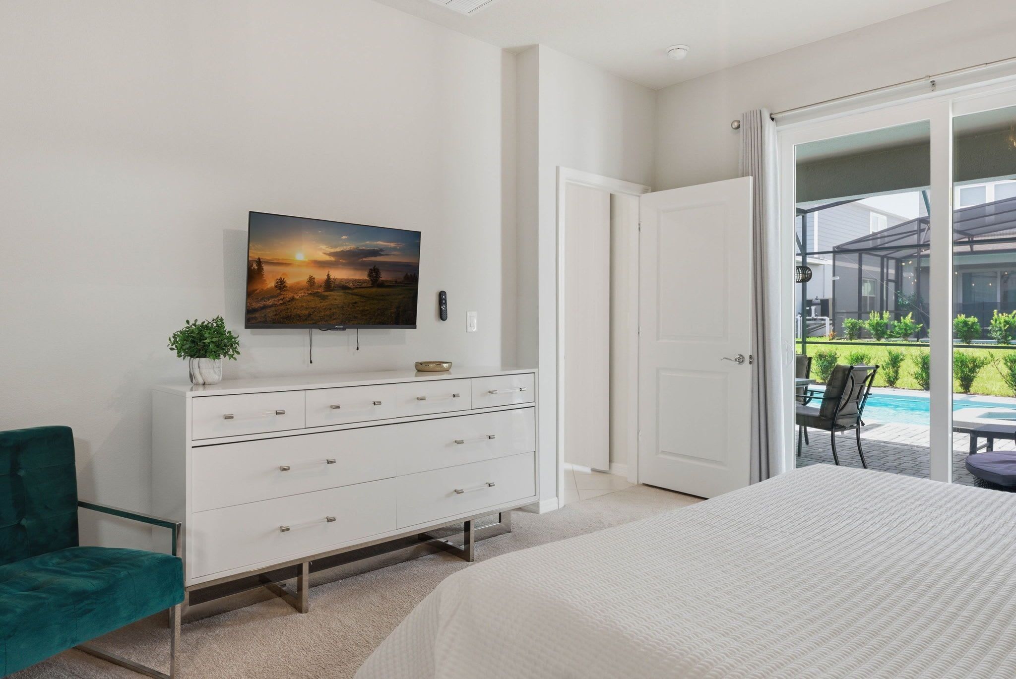 A bedroom with a bed, a dresser, a TV on the wall, and a sliding glass door leading to a balcony with a pool.