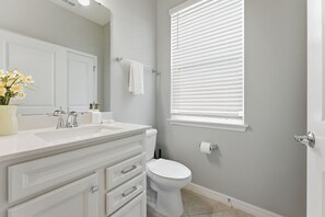 Un bagno con lavandino bianco, specchio e WC. Si vede una finestra con persiane e un asciugamano su un portasciugamani.