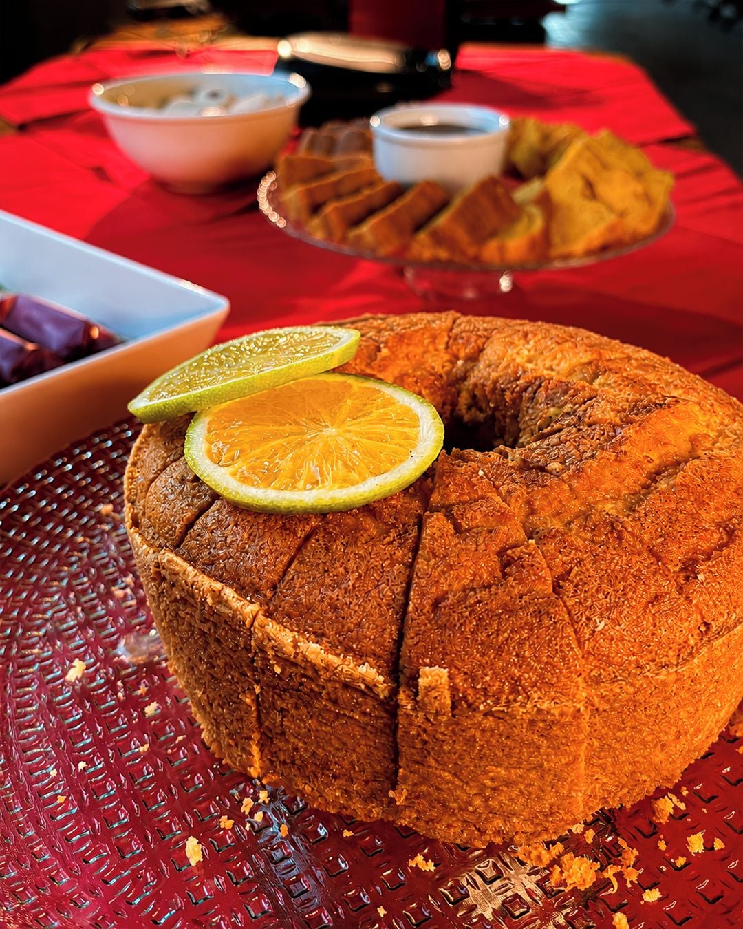 A bundt cake with lemon slices on a textured plate, with bowls and a red tablecloth in the background.