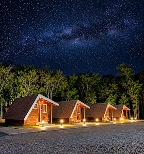 A row of wooden cabins with pitched roofs, set against a night sky with visible stars and the Milky Way.