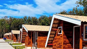 A row of wooden cabins with a tiled roof, each with a door and window, set against a backdrop of trees and a clear sky. - Chalés Eco Thermas (Tupã)