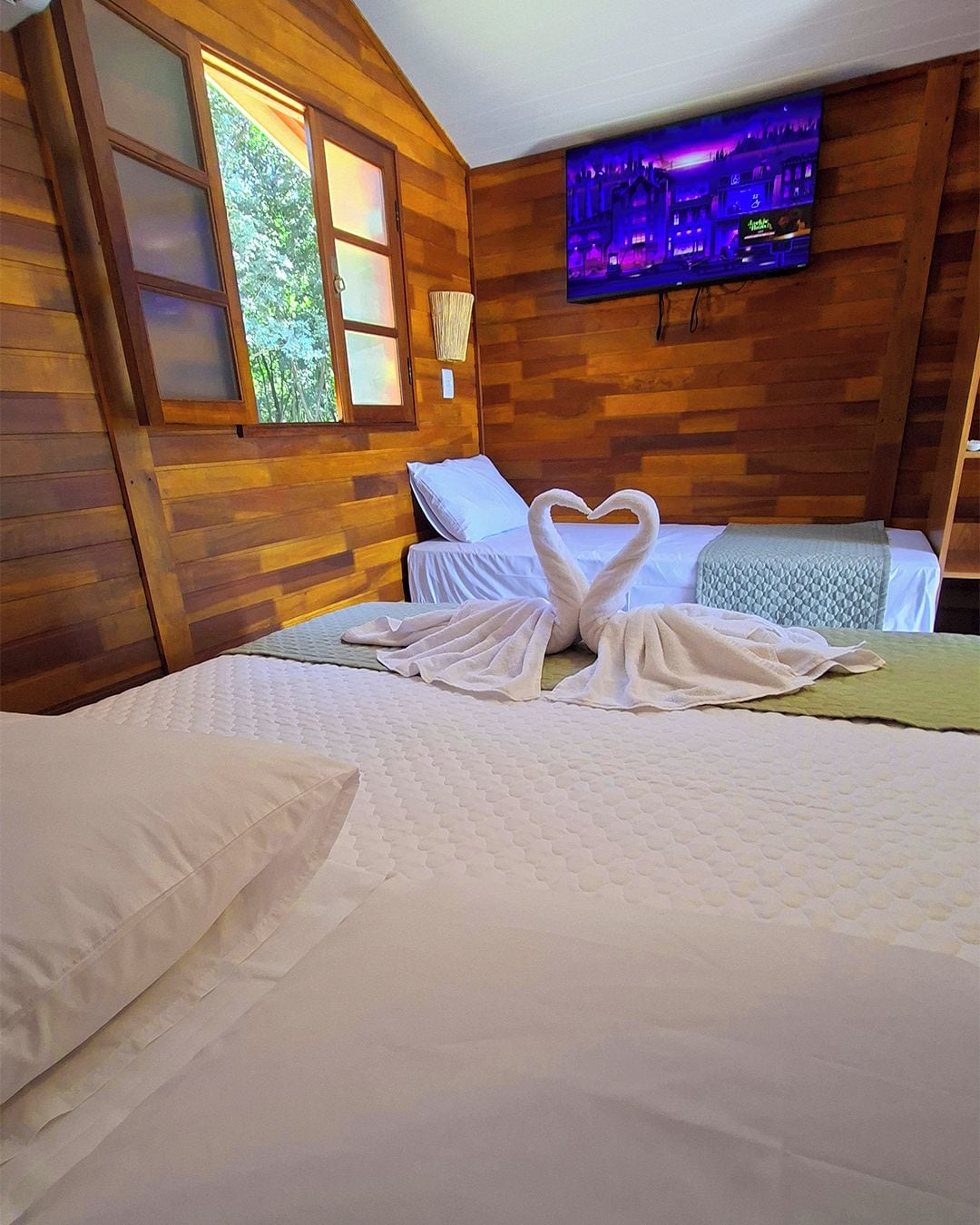 Une chambre d’hôtel avec un décor intérieur en bois, un lit avec une literie blanche, une disposition de serviettes en forme de cœur et un téléviseur fixé au mur.
