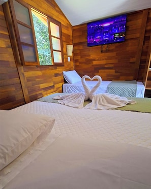 Habitación de hotel con detalles de madera, cama con ropa blanca, toallas dispuestas en forma de corazón y un televisor montado en la pared.