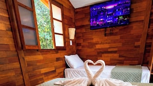 A room with wooden walls, a bed with two pillows, a heart-shaped towel arrangement, a window with a view of greenery, and a television mounted on the wall. - Chalés Eco Thermas (Tupã)