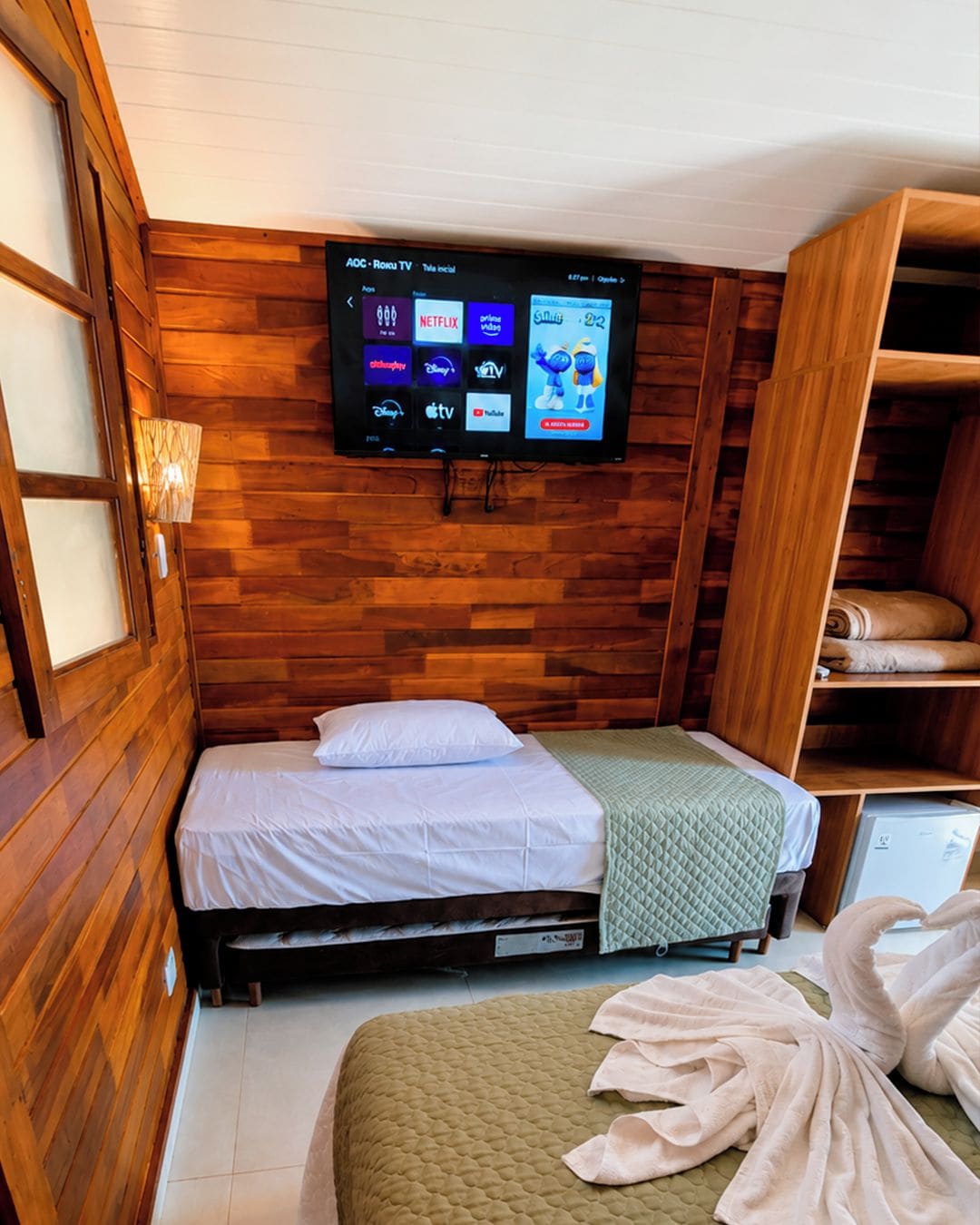 A hotel room with a bed, a television mounted on the wall, a wooden wardrobe, and a small table.