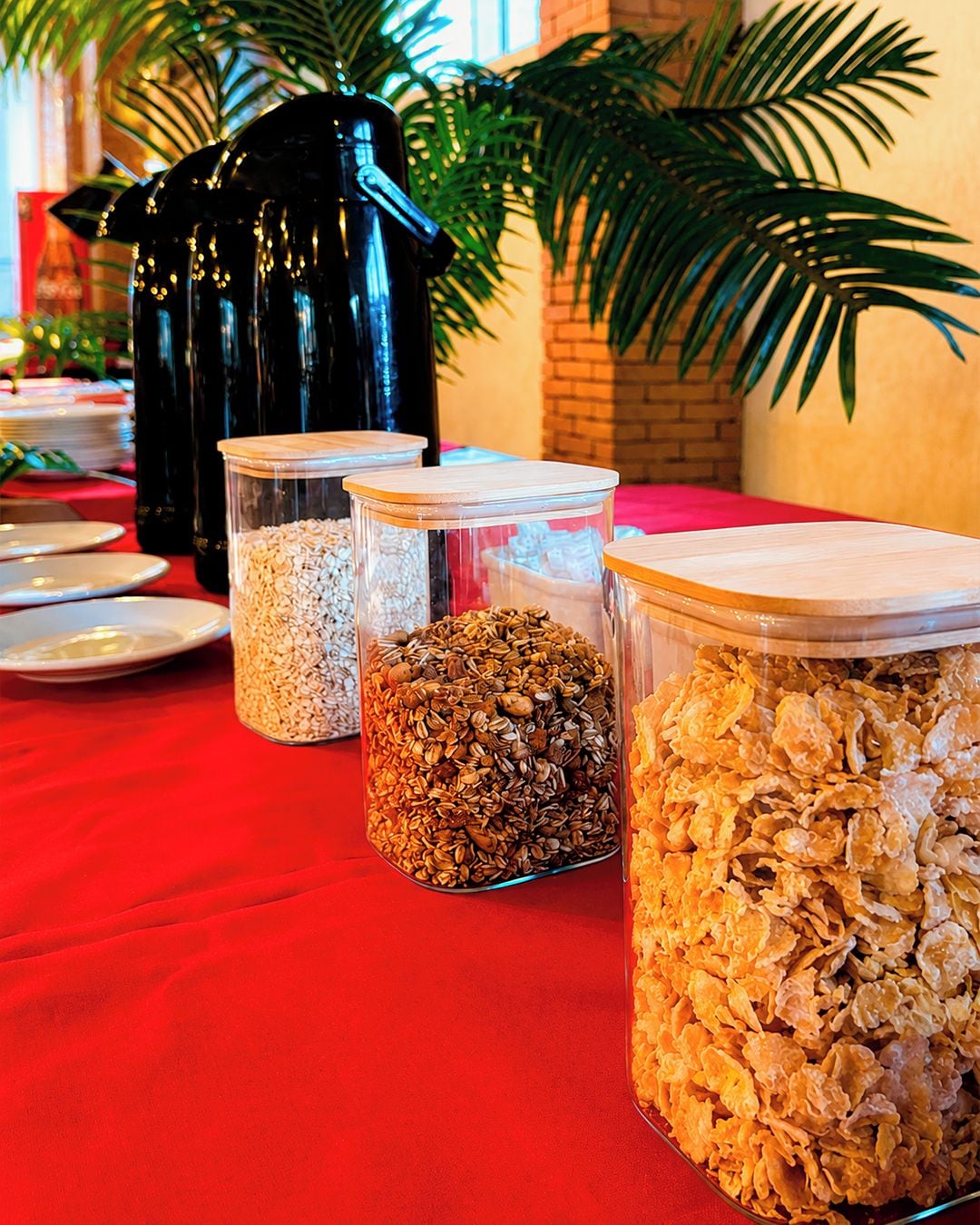 A table with three glass containers of different food items, a black coffee pot, and a red tablecloth.