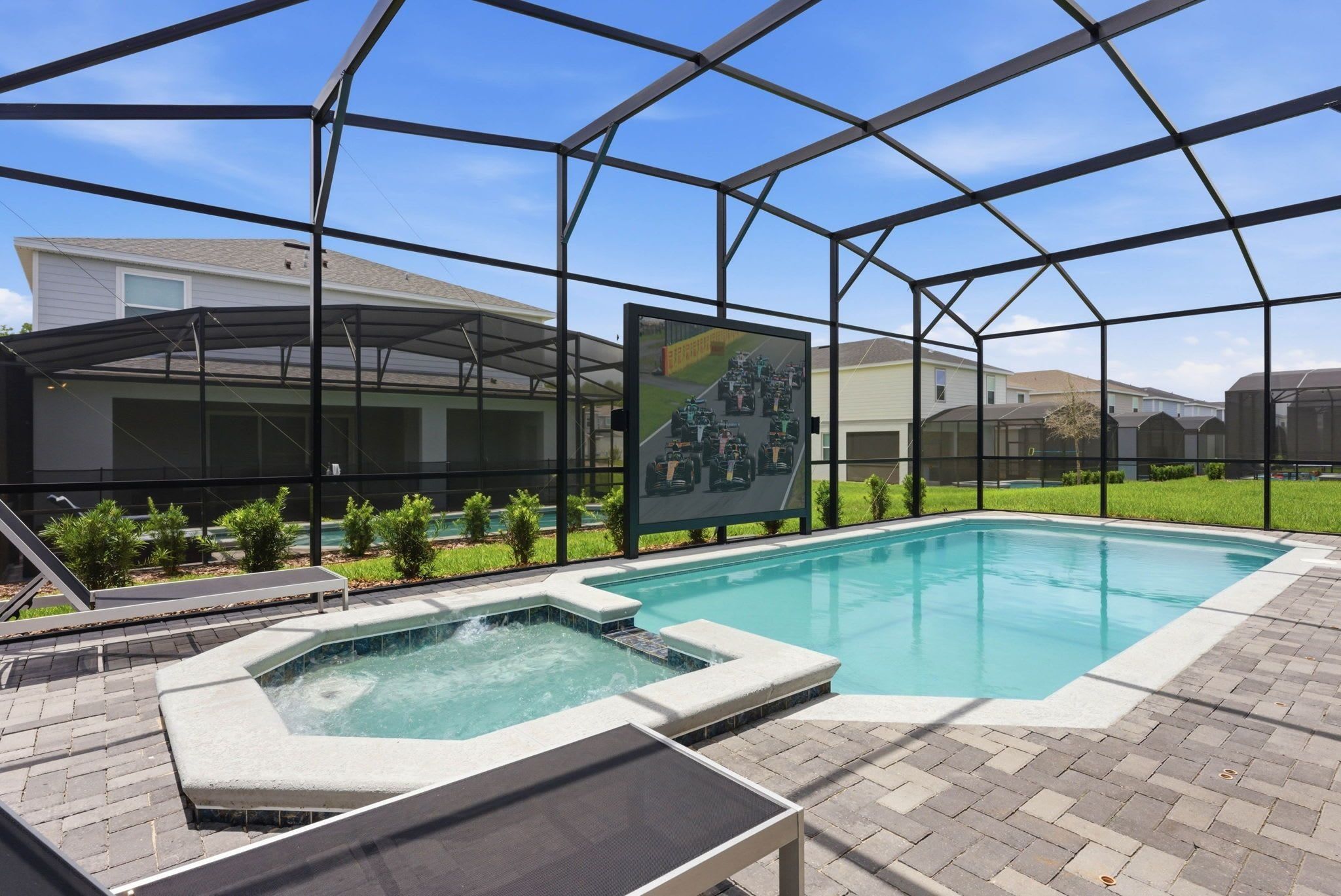 A swimming pool with a hot tub, surrounded by a covered patio area with a large screen displaying a racing event.