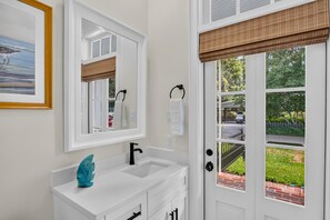 Une salle de bain avec un meuble-lavabo blanc, un miroir et une fenêtre donnant sur une rue et des espaces verts.