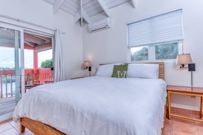 Ein Schlafzimmer mit einem großen Bett, zwei Nachttischen, einem Fenster mit Jalousien, einer Klimaanlage und einem Balkon mit roter Brüstung.