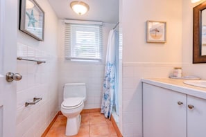 Ein Badezimmer mit weißen Fliesen, einer Toilette, einer Duschvorhang, einem Fenster mit Jalousien und einem Waschbecken mit Spiegel darüber.