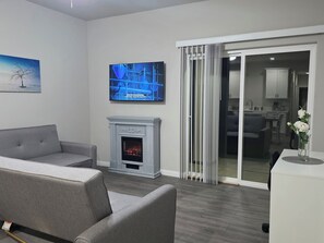 Ein modernes Wohnzimmer mit Kamin, einer grauen Couch, einem Flachbildfernseher und einer Glastür, die in eine Küche führt.