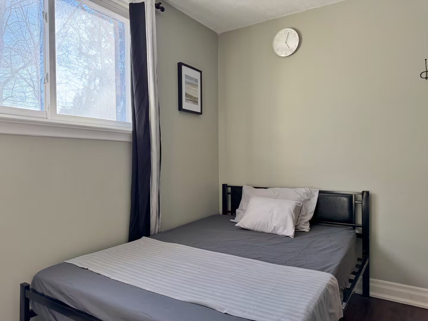 A bedroom with a single bed, a window with curtains, a wall clock, and a framed picture.