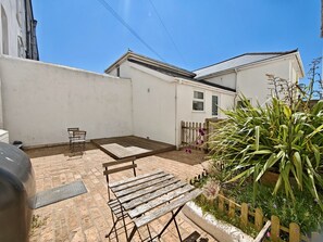 A patio with a wooden table and chairs, a barbecue grill, and a garden with plants and a wooden fence. - Private Beach-Front Apartment on the English Riviera with outdoor courtyard (Paignton)