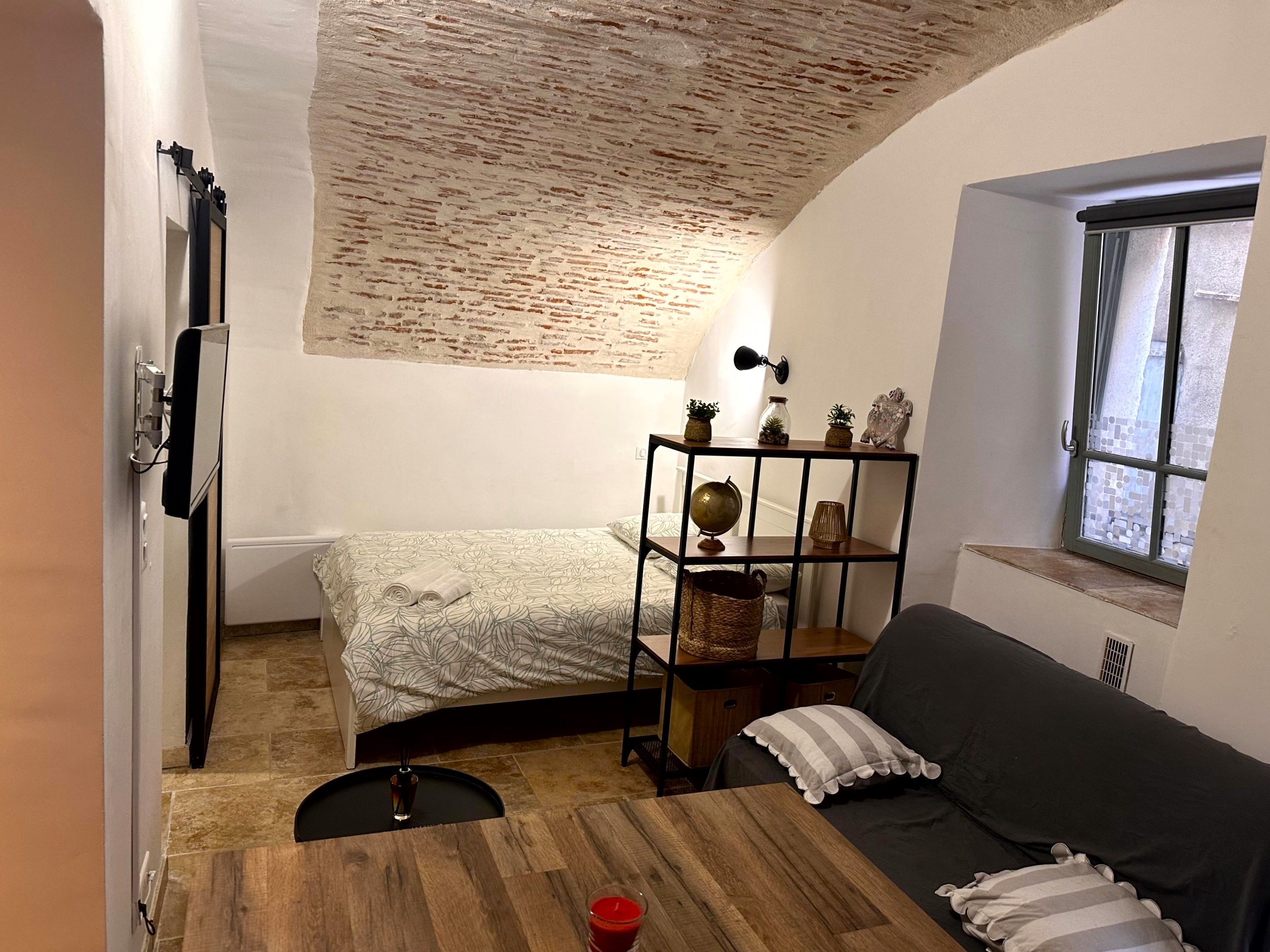 Une pièce de taille modeste comprenant un lit, une petite table, un canapé, une étagère avec des objets décoratifs et une fenêtre donnant sur un paysage agréable.