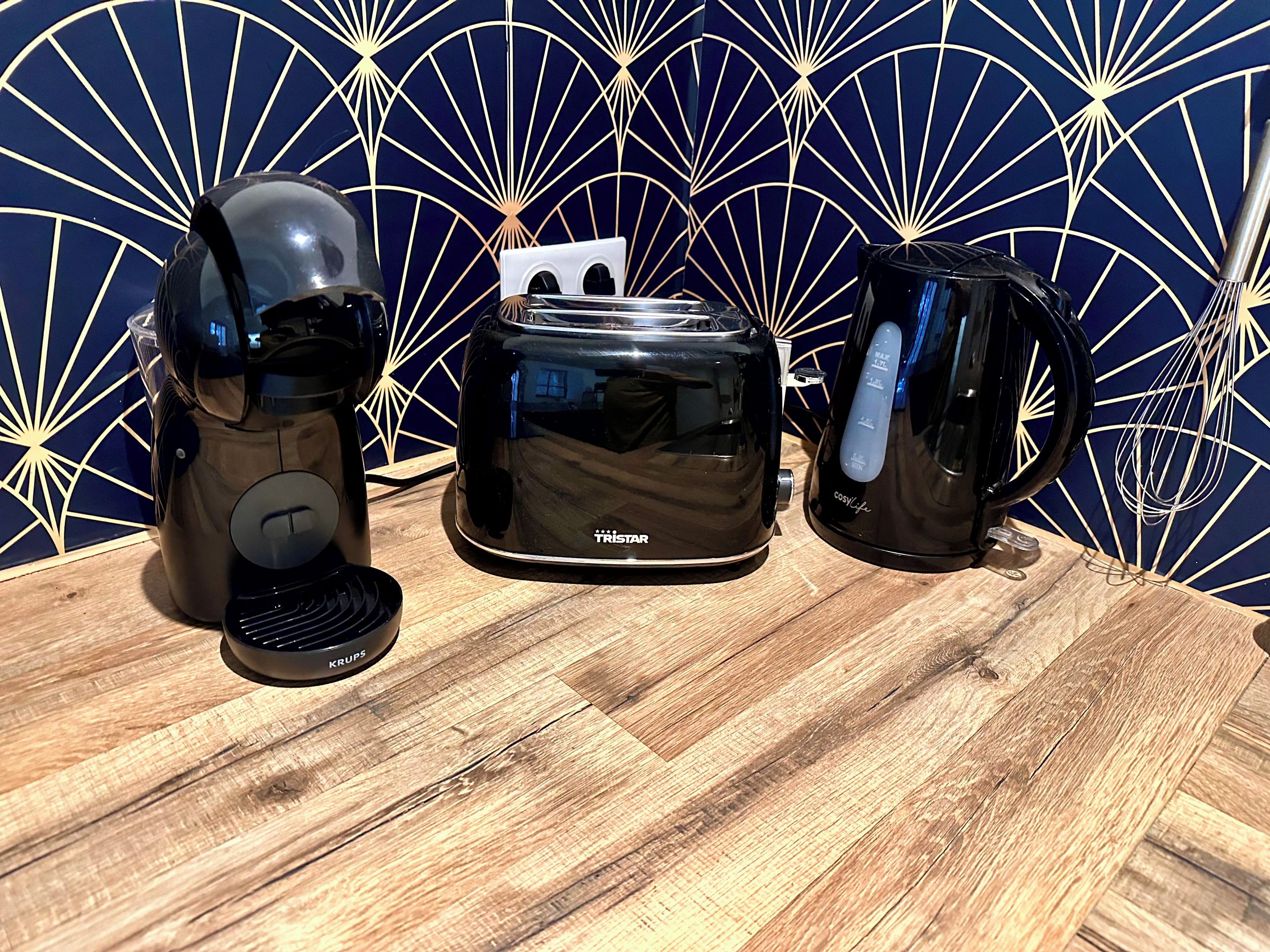 A coffee maker, a toaster, and a mug on a wooden table with a geometric patterned wall in the background.