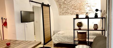 A compact room with a bed, a sofa, a small table, and a TV mounted on the wall.