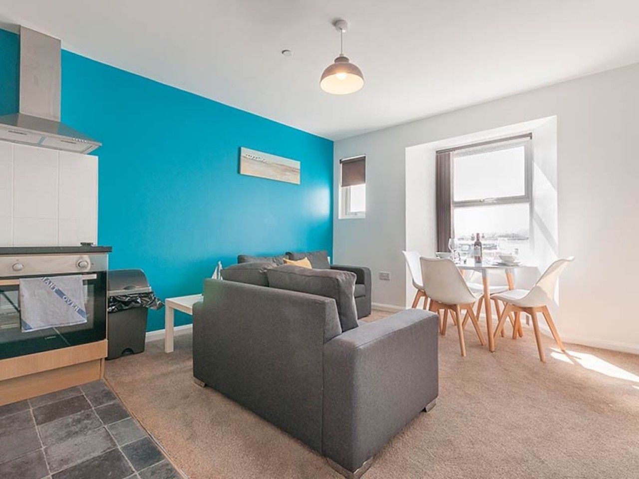Ein modernes Wohnzimmer mit blauer Akzentwand, ein Essbereich mit weißen Stühlen und eine Küche mit Einbauofen.