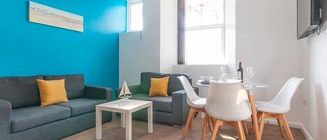 Una sala de estar moderna con una pared azul de acento, un sofá gris, una mesa de centro blanca y un comedor con sillas blancas y una mesita.
