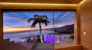 Ein modernes Interieur mit einem großen Panoramafenster, das auf einen Küsten-Sonnenuntergang, eine Palme und eine Treppe zu einem Außenbereich blickt.