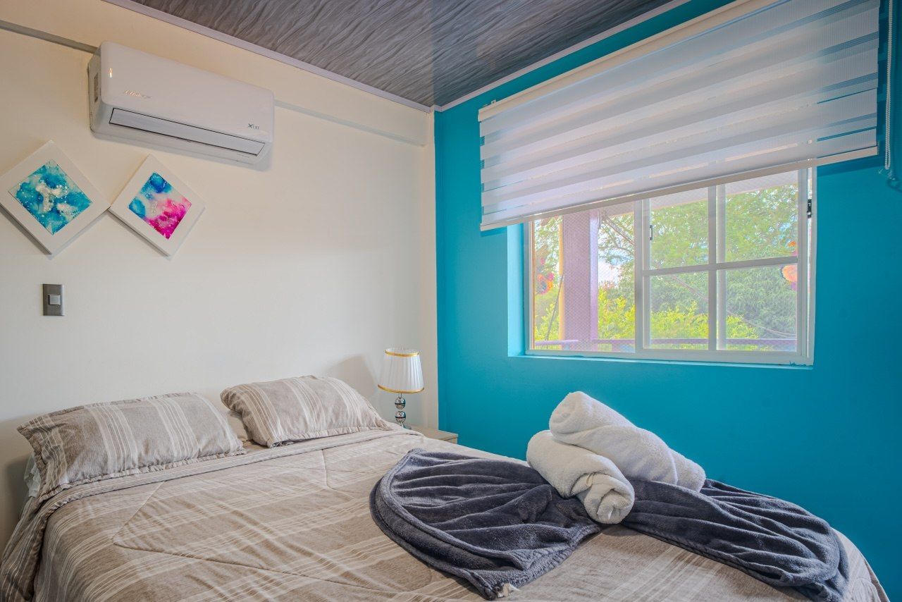 Un dormitorio con una cama, una ventana con persianas, un aire acondicionado y dos obras de arte colgadas en la pared.