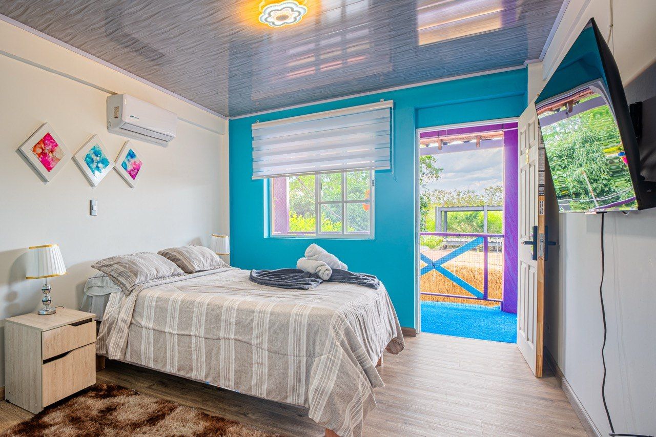 A bedroom with a bed, bedside table, air conditioning unit, window with blinds, and a view of an outdoor area.