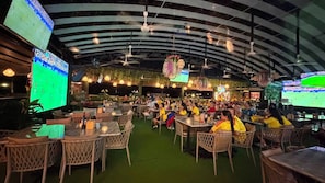 A crowded indoor dining area with tables and chairs, large screens displaying sports events, and hanging floral decorations.