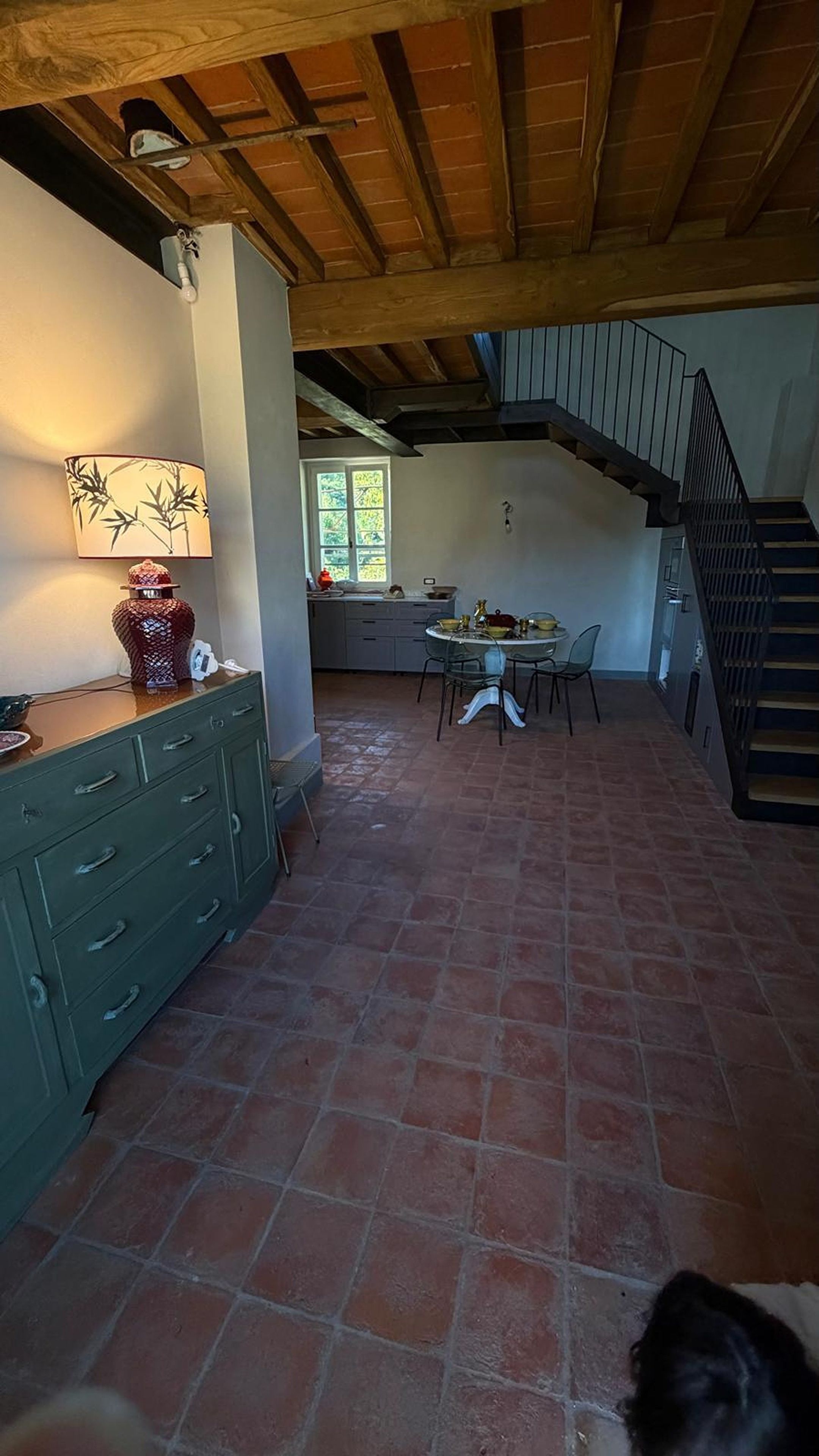 Ein geräumiger Raum mit hoher Decke und freiliegenden Holzbalken, ein Essbereich mit Tisch und Stühlen sowie eine Treppe, die zu einer oberen Ebene führt.