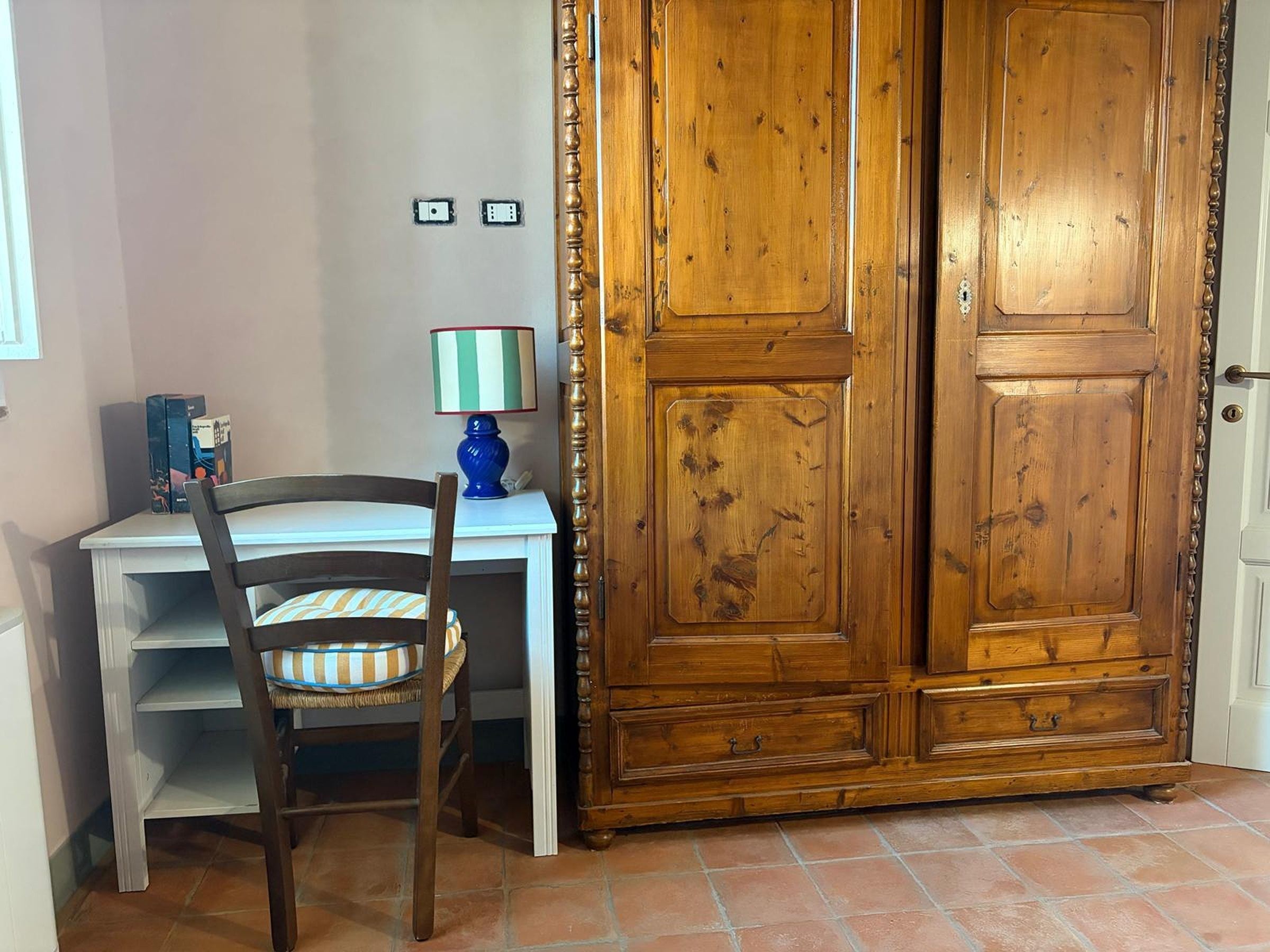 A room with a wooden wardrobe, a desk with a chair, and a lamp.