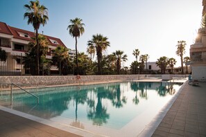 A swimming pool with a stone wall and palm trees, surrounded by buildings with red roofs. - DANICO PLAYA HONDA 51/C (Playa de las Américas)