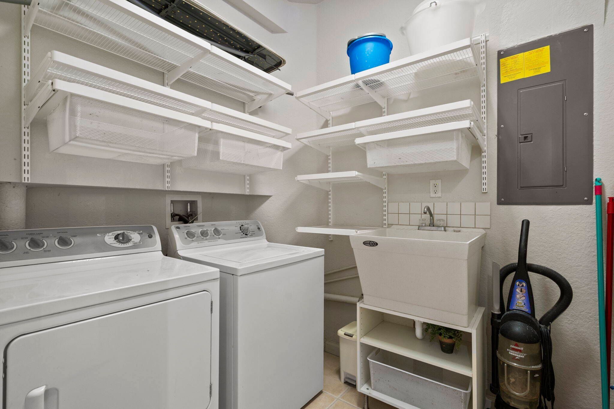 A laundry room with two washing machines, shelves for storage, and a vacuum cleaner.