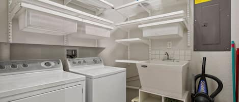 A laundry room with two washing machines, shelves for storage, and a vacuum cleaner.