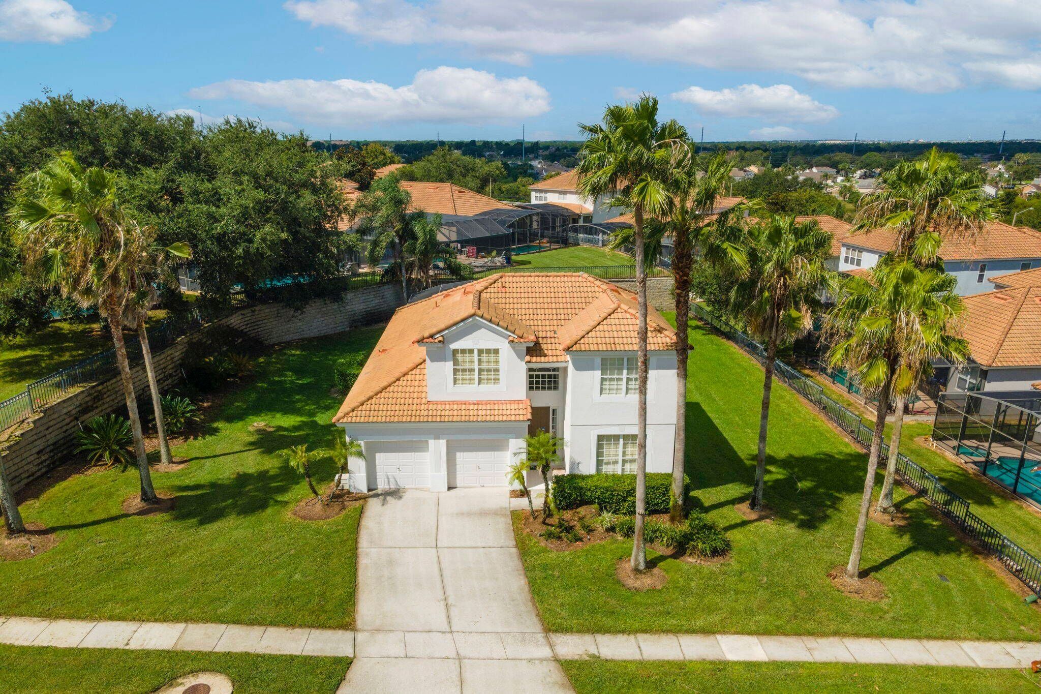 A residential area with a white house featuring a tiled roof, surrounded by palm trees and a well-maintained lawn.