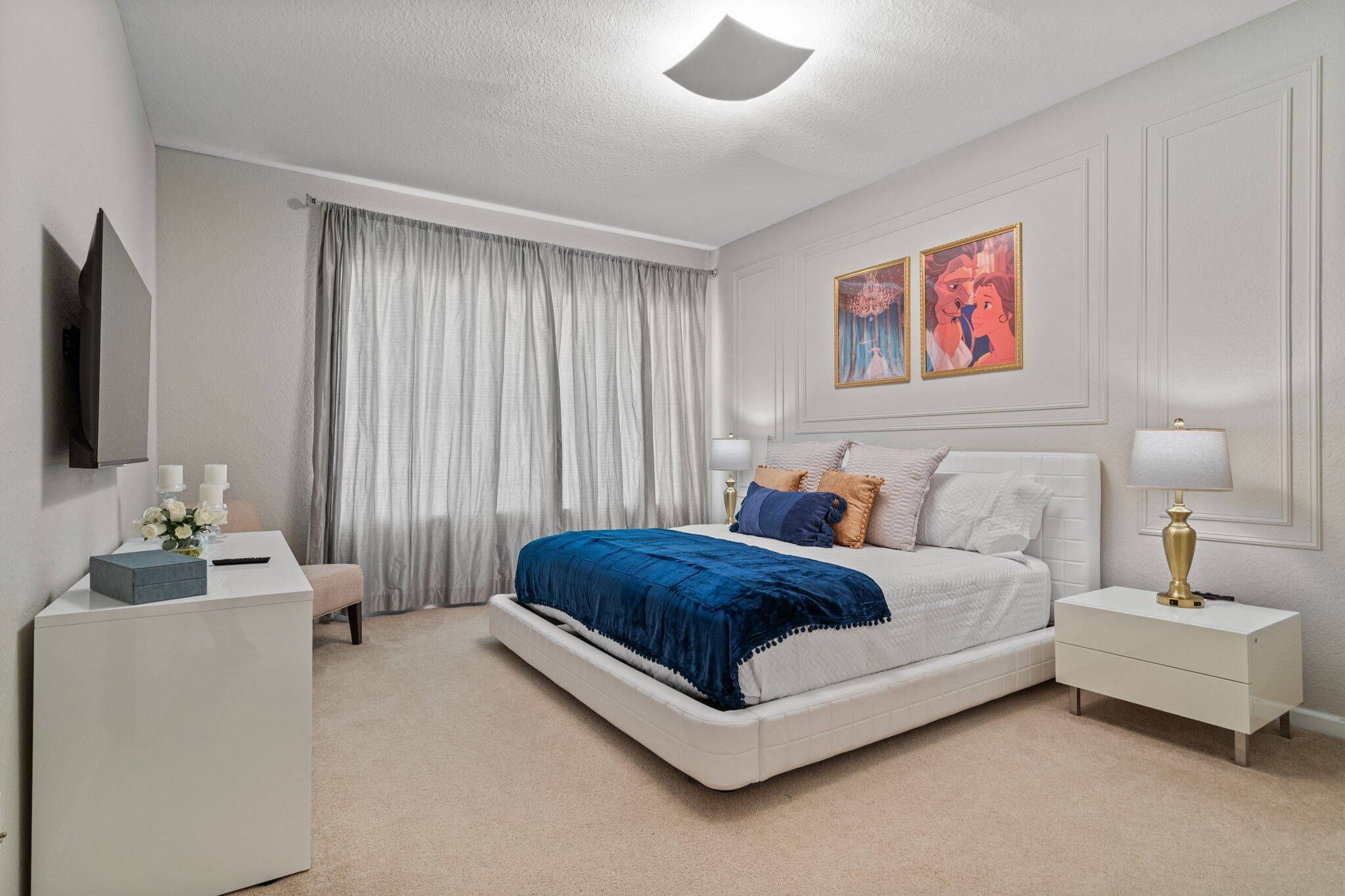 Un dormitorio con cama, mesitas de noche, escritorio, silla, televisor y dos cuadros en la pared.