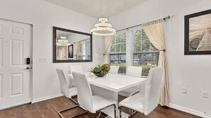 Ein Esszimmer mit weißem Tisch, vier Stühlen, einer Chandelierleuchtre, einem fenster mit Vorhängen und einem Spiegel an der Wand.