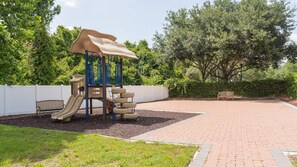 Ein Spielplatz mit Rutsche und Klettergerüst, umgeben von einem Pflasterweg und Grünflächen.