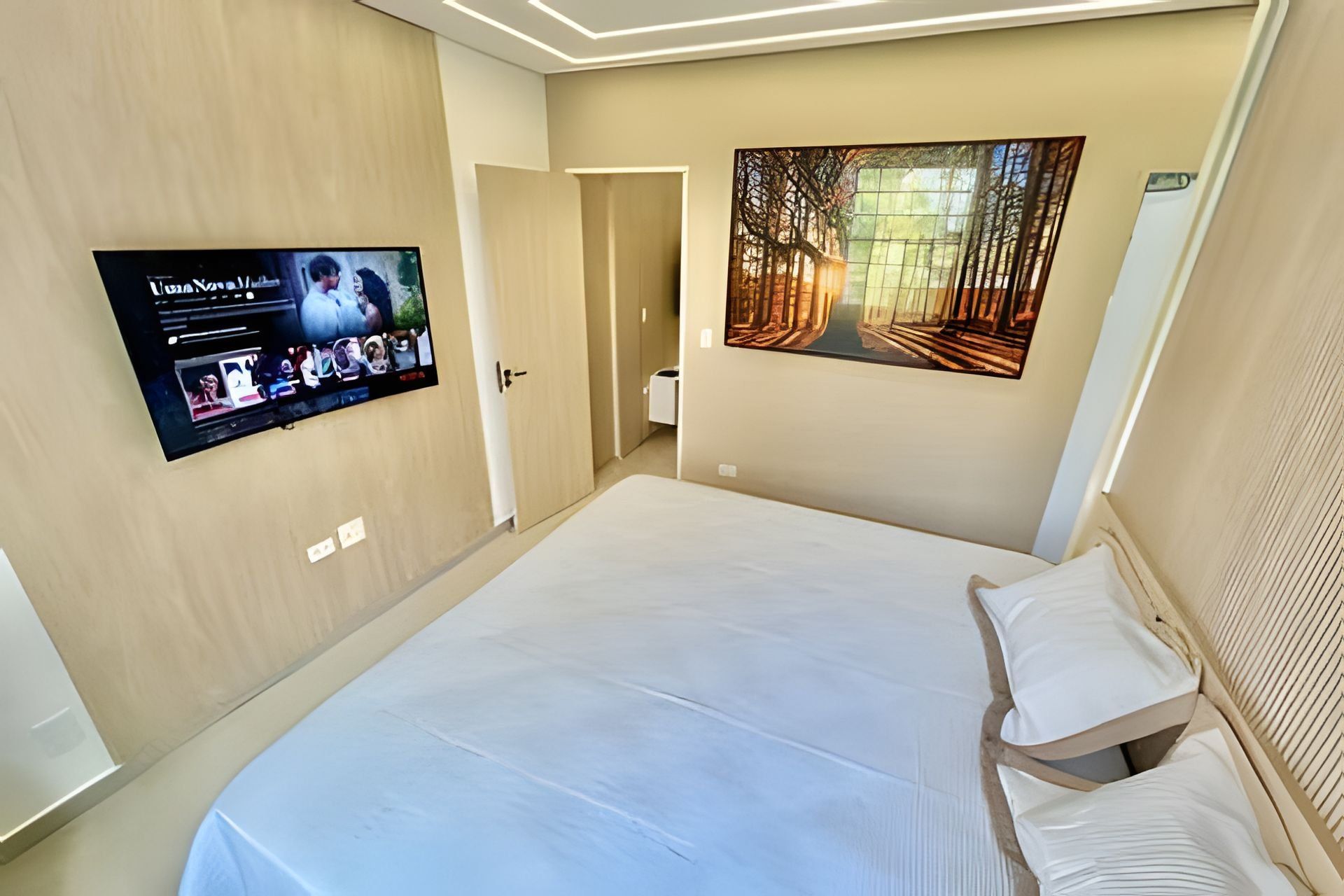 Habitación de hotel con una cama grande, un televisor montado que muestra un noticiero y un cuadro con una escena de bosque.