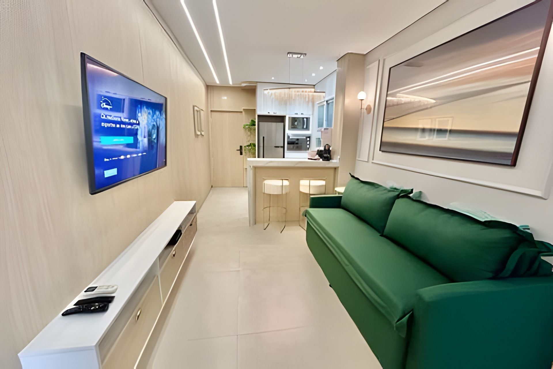 Una sala de estar moderna con un televisor de pantalla plana montado en la pared, un sofá verde, una mesa consola blanca y una zona de cocina al fondo.