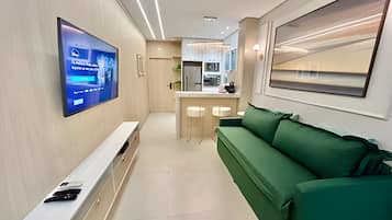 Una sala de estar moderna con un televisor de pantalla plana montado en la pared, un sofá verde, una mesa consola blanca y una zona de cocina al fondo.