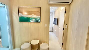Un pasillo con un cuadro de un paisaje montañoso, un televisor montado en la pared y un pequeño rincón de descanso con tres sillas.