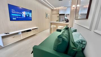 A modern living room with a flat-screen TV mounted on the wall, a white entertainment unit, a green sofa with cushions, and a kitchen area in the background.