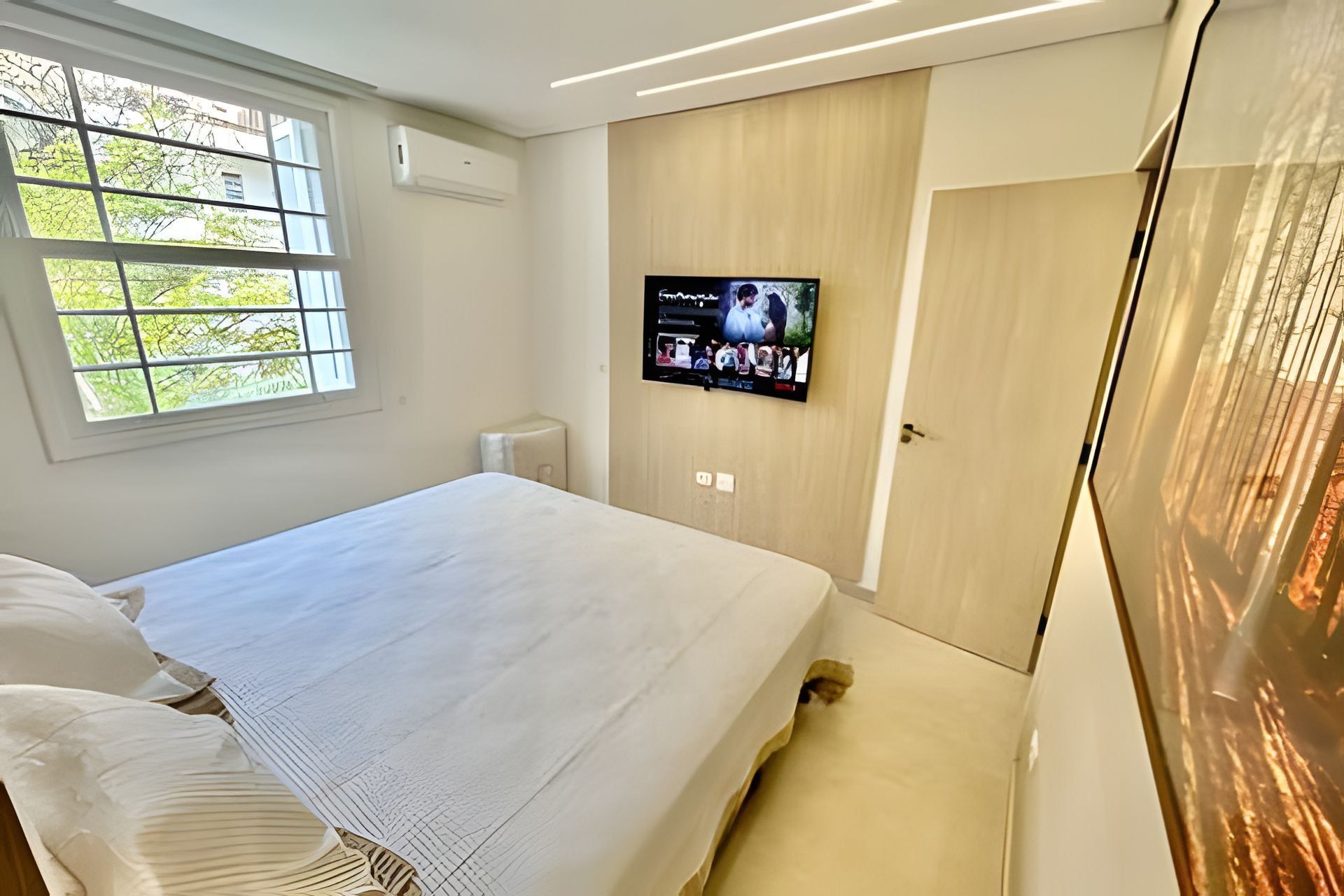 A bedroom with a bed, a TV mounted on the wall, and a window with a view of trees.