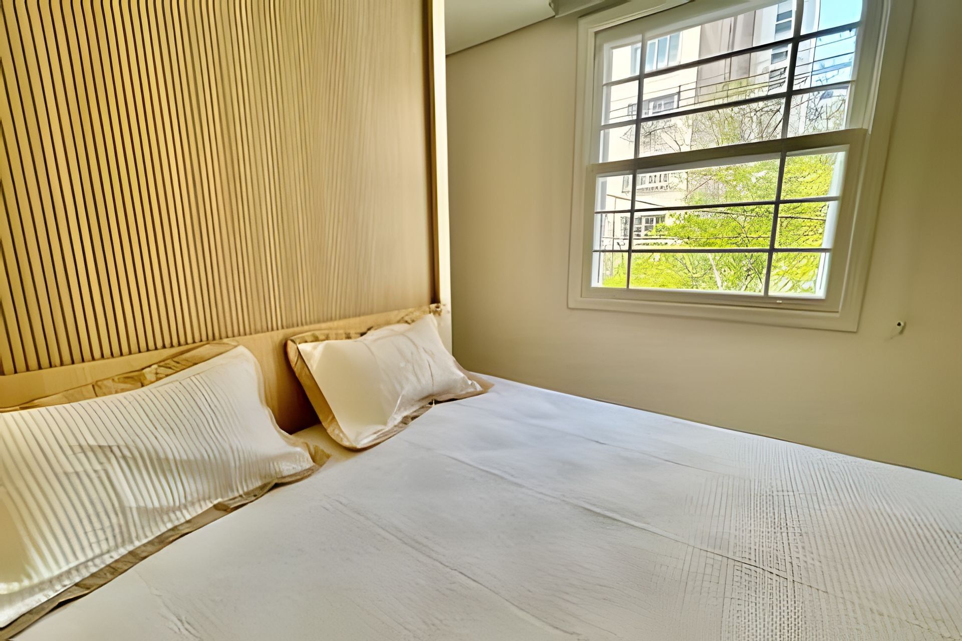 A bedroom with a large bed, two pillows, a wooden headboard, and a window with a view of greenery.