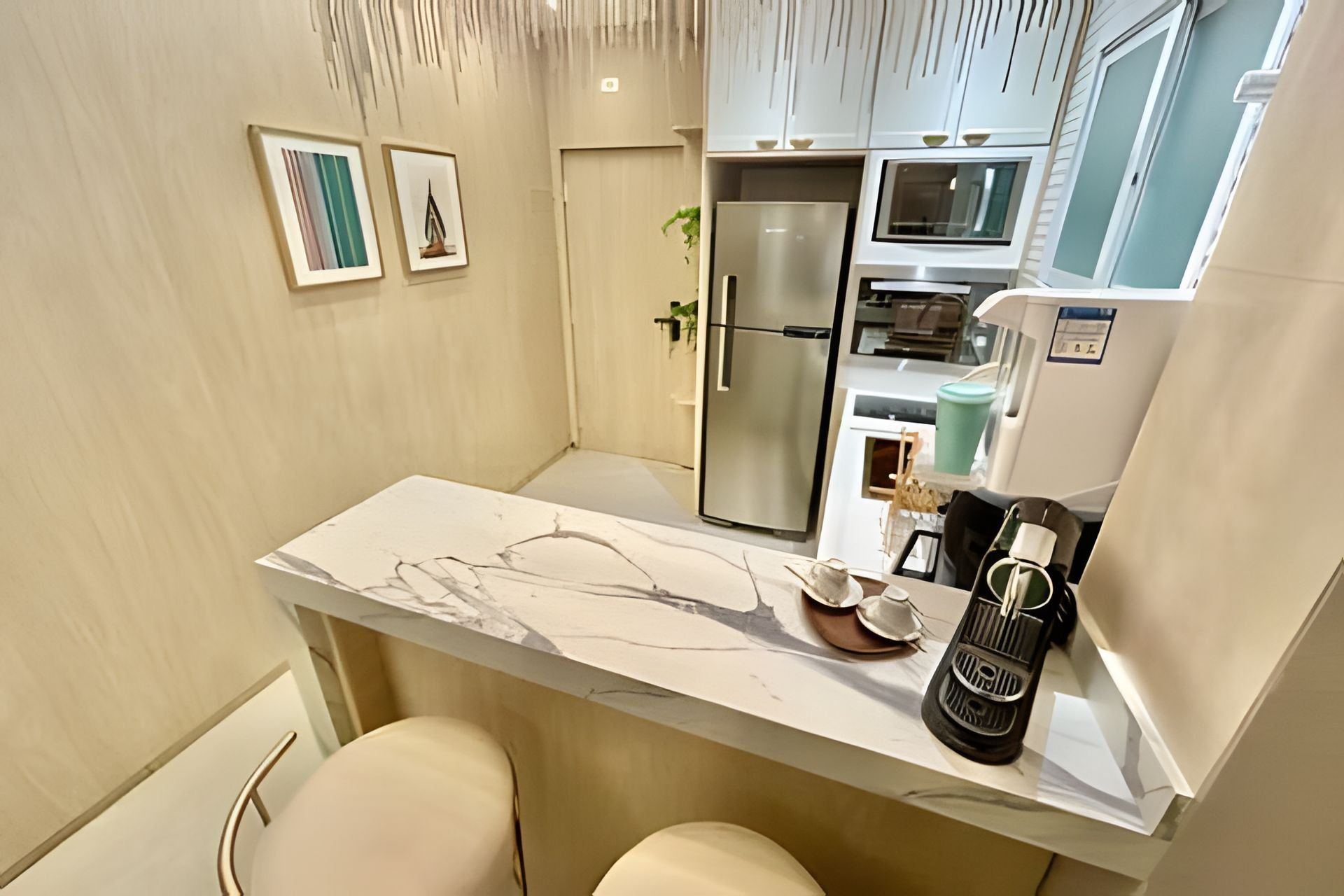 Cocina compacta con encimera de mármol, refrigerador y lavamanos. Hay dos taburetes y una mesita con una botella y una taza.