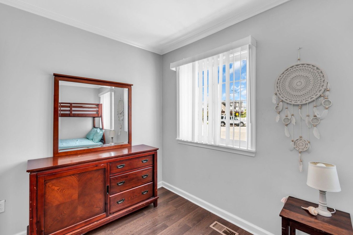 A room with a wooden dresser, a mirror, a window with blinds, and a dreamcatcher on the wall.
